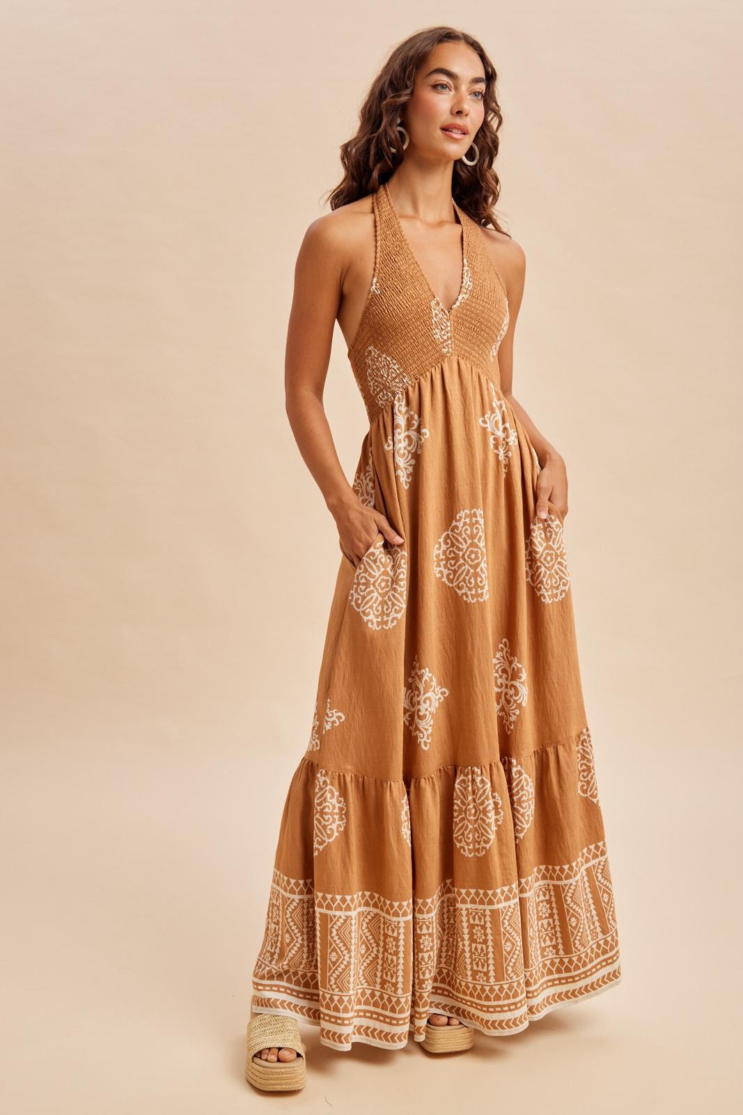 woman wearing a brown maxi dress with white intricate patterns and pockets, standing against a neutral background