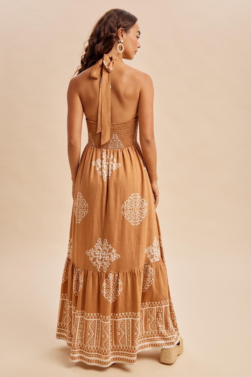 woman wearing a backless tan maxi dress with white embroidered patterns and a halter tie detail