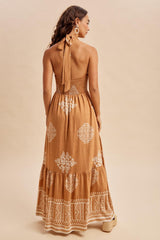woman wearing a backless tan maxi dress with white embroidered patterns and a halter tie detail