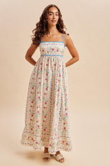 woman wearing a floral maxi dress with thin blue straps and a fitted bodice, standing with hands on hips