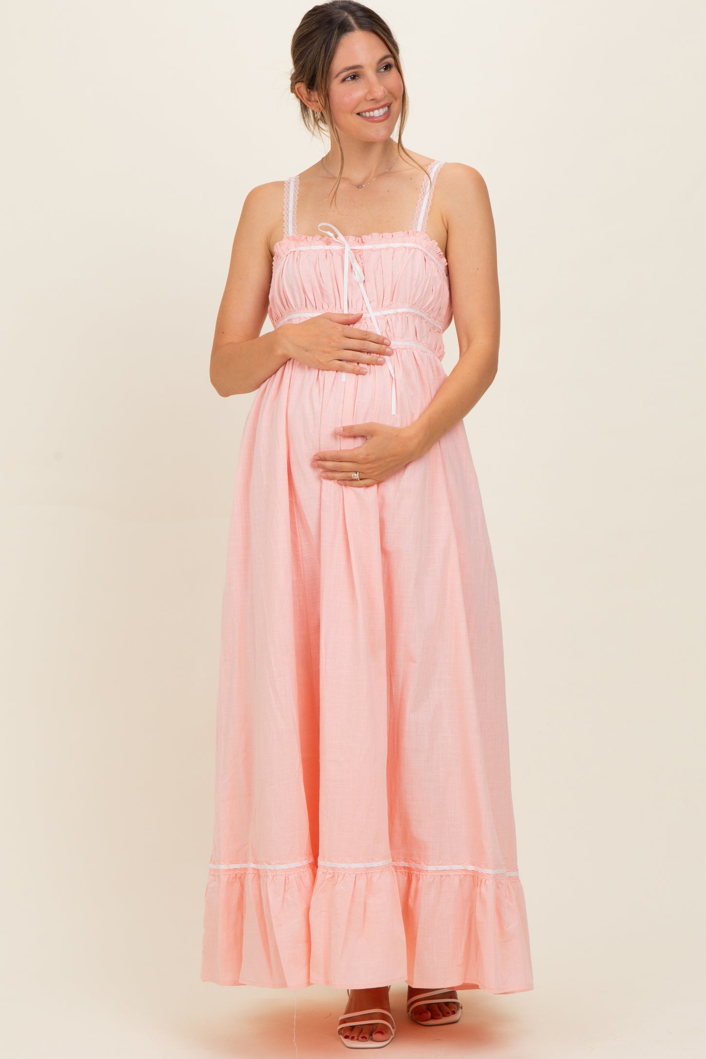 woman wearing a light pink maternity maxi dress with ruffle hem and adjustable straps, gently holding her baby bump