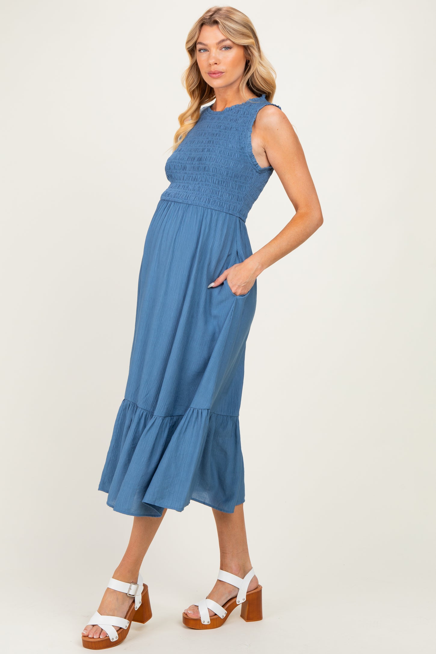 woman wearing a blue smocked maternity dress with a tiered skirt and pockets, showcasing her baby bump