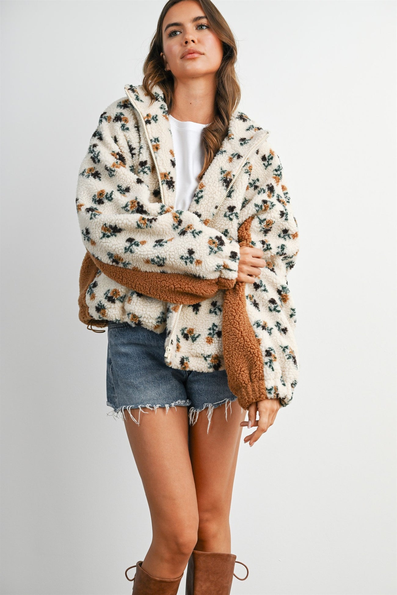 woman wearing a cream floral patterned fuzzy jacket with brown accents, paired with denim shorts and tall brown boots