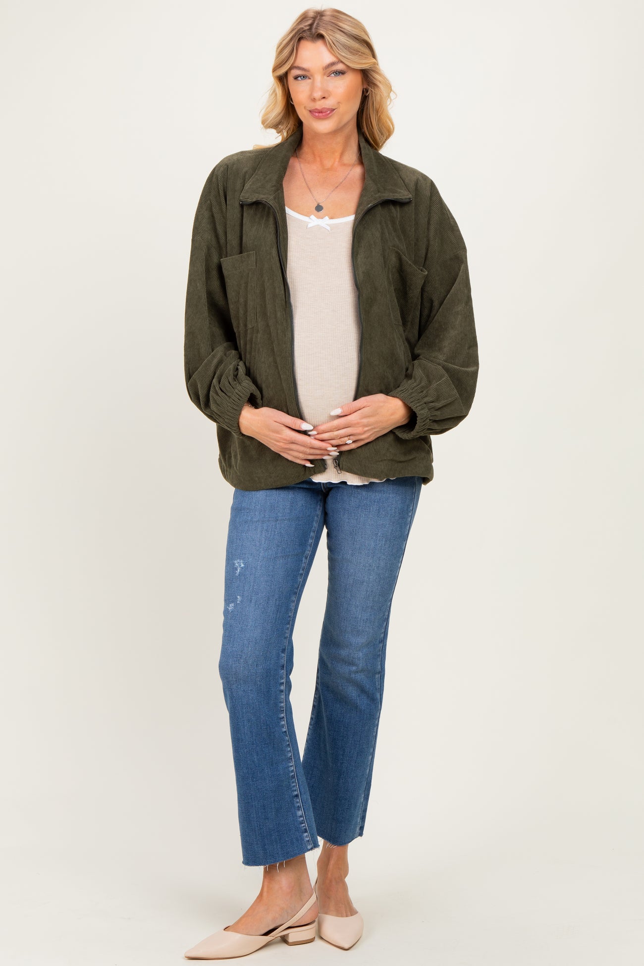 woman wearing a green corduroy maternity jacket over a beige top and holding her baby bump with blue jeans and beige flats