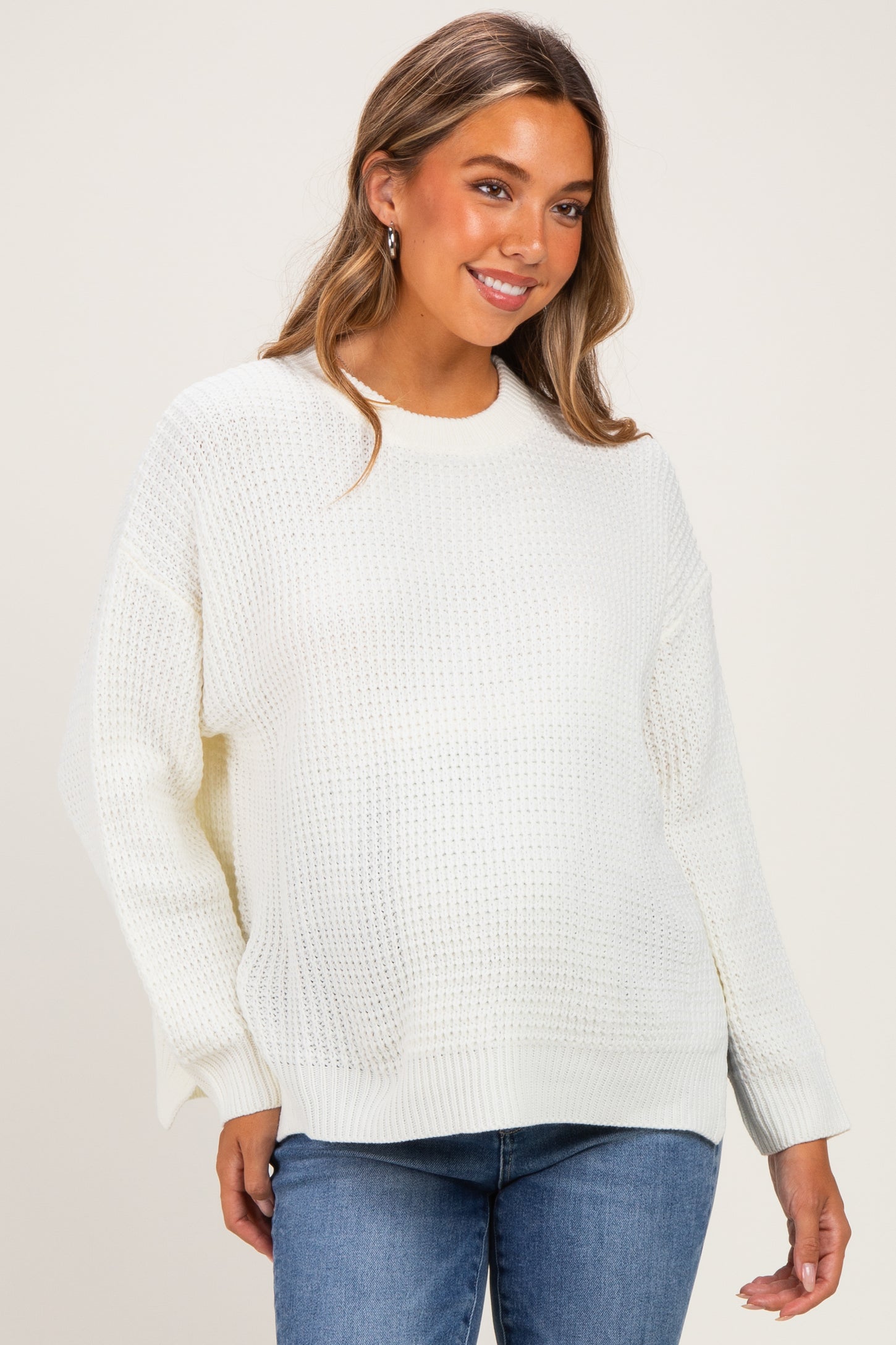 woman wearing a white knitted maternity sweater with a relaxed fit and side slits, paired with blue jeans and smiling