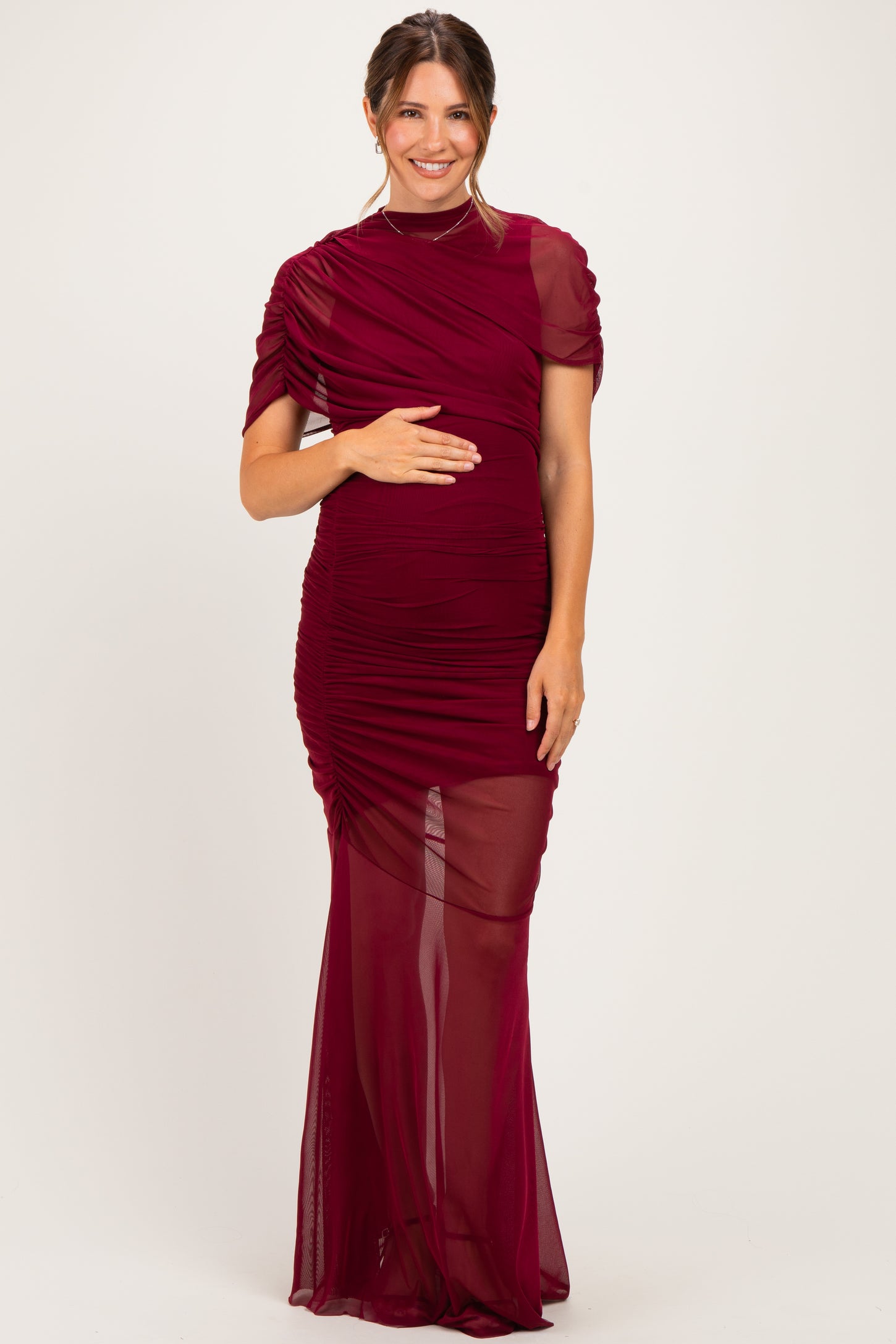 woman wearing a burgundy maternity gown with sheer sleeves, holding her baby bump and featuring ruched details