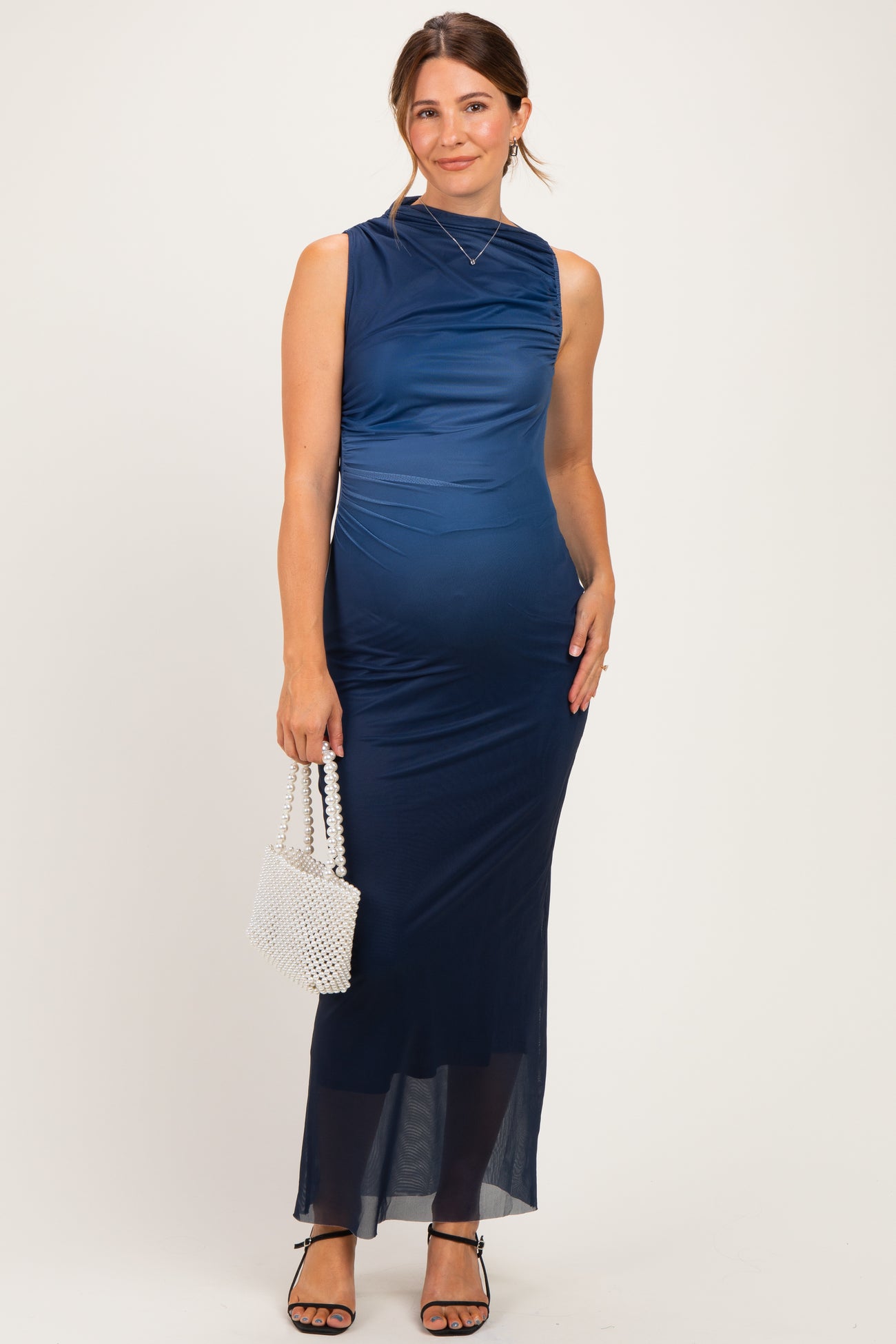 woman wearing a navy blue sleeveless maternity dress with ruching and a sheer bottom, holding a small white bag