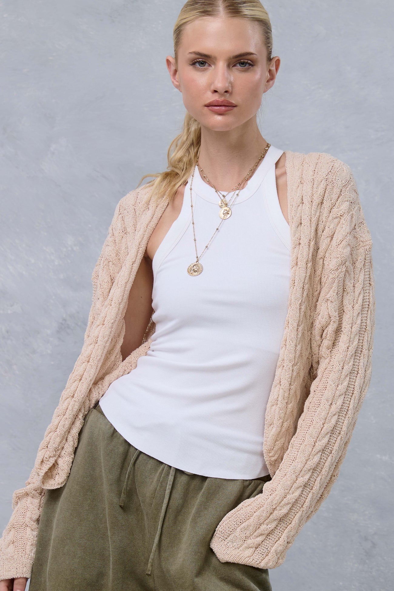 woman wearing a beige cable-knit cardigan over a white tank top and olive green pants, styled with layered necklaces