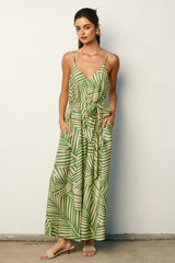woman wearing a green palm leaf patterned maxi dress with a v-neckline and adjustable tie waist, standing against a wall