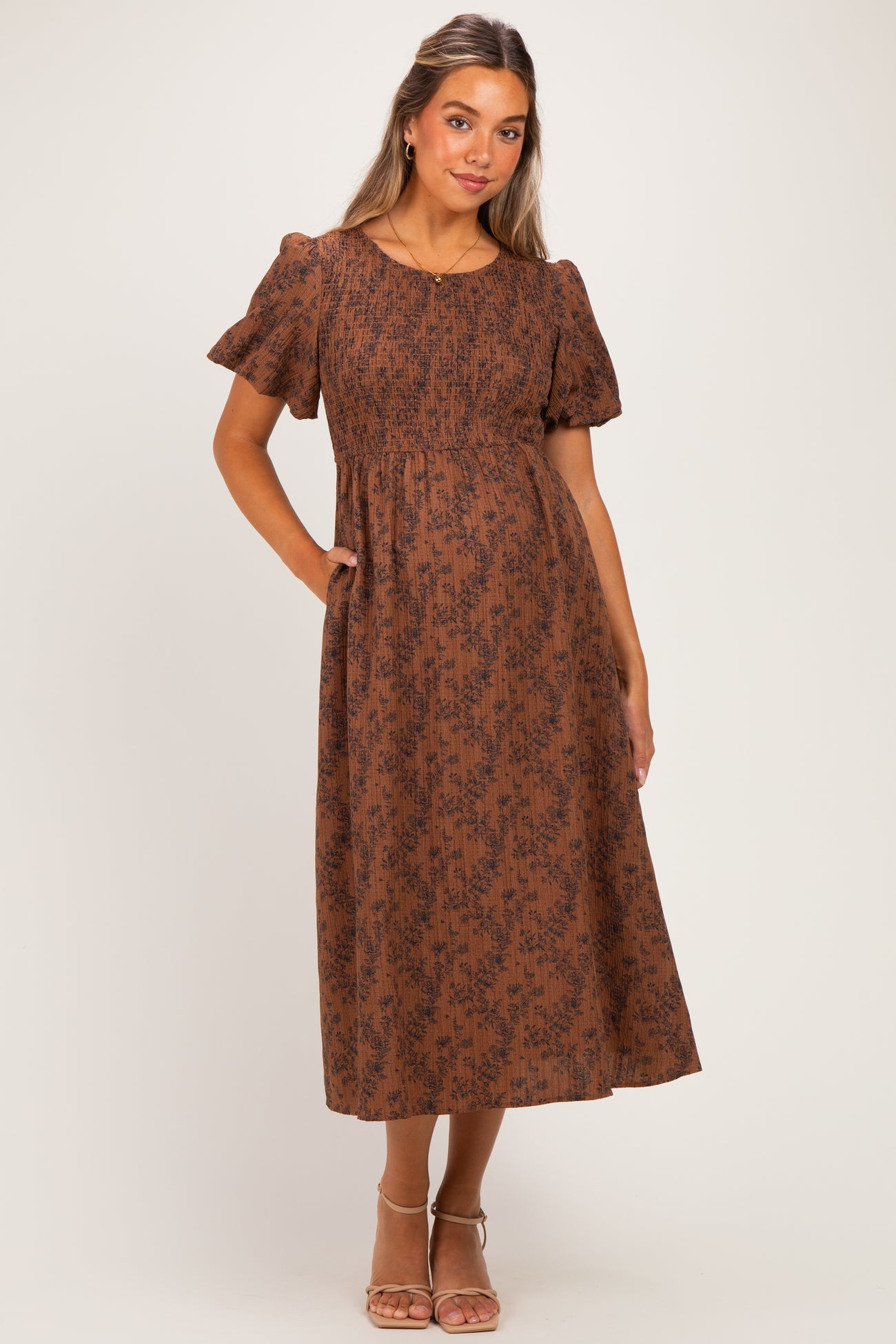 woman wearing a brown floral print maternity dress with short puff sleeves and a smocked bodice, standing with hands in pockets