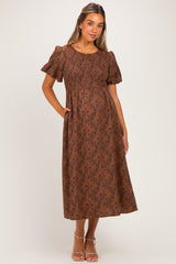 woman wearing a brown floral print maternity dress with short puff sleeves and a smocked bodice, standing with hands in pockets