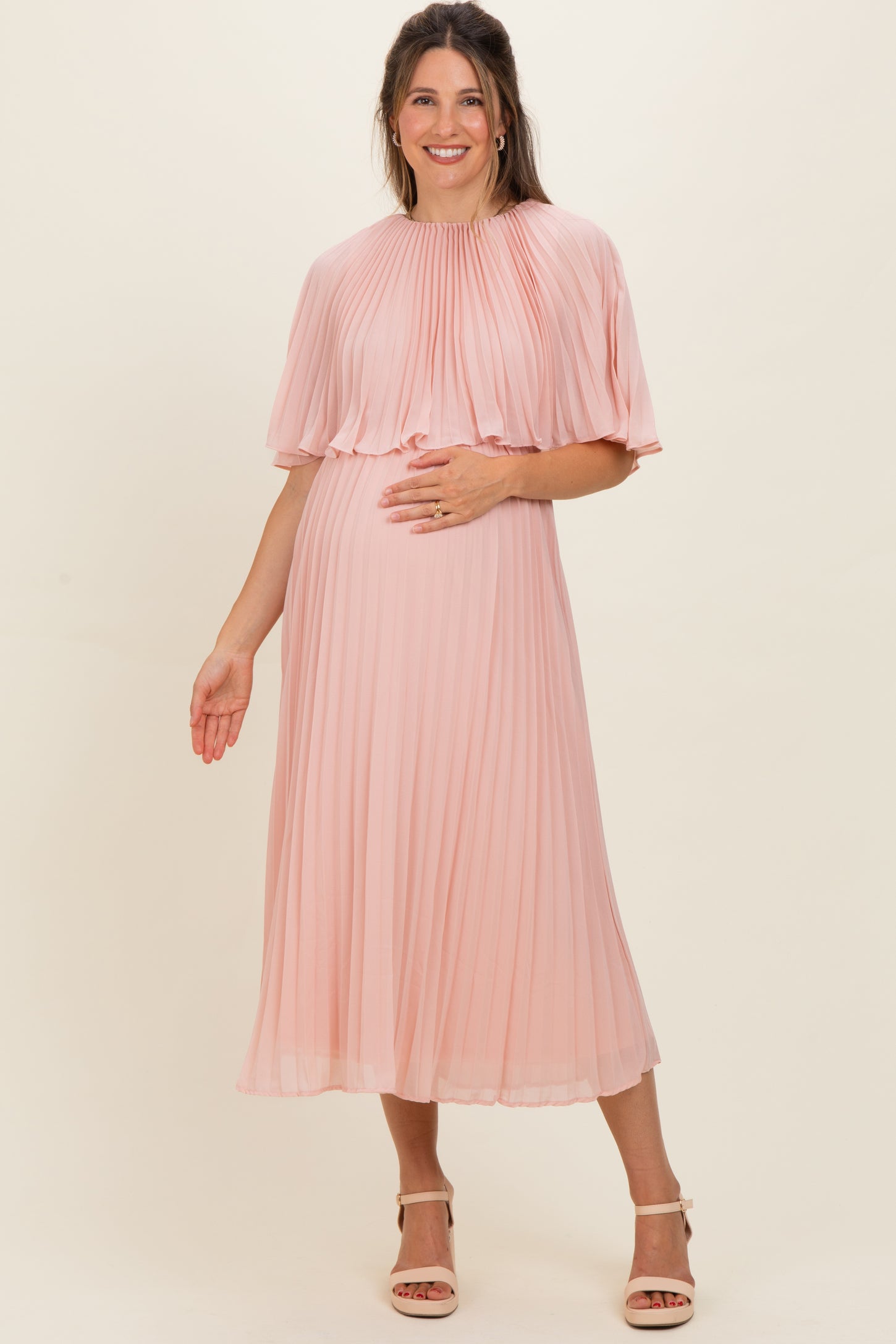 woman wearing a blush pink pleated maternity dress with a flowing cape and holding her baby bump