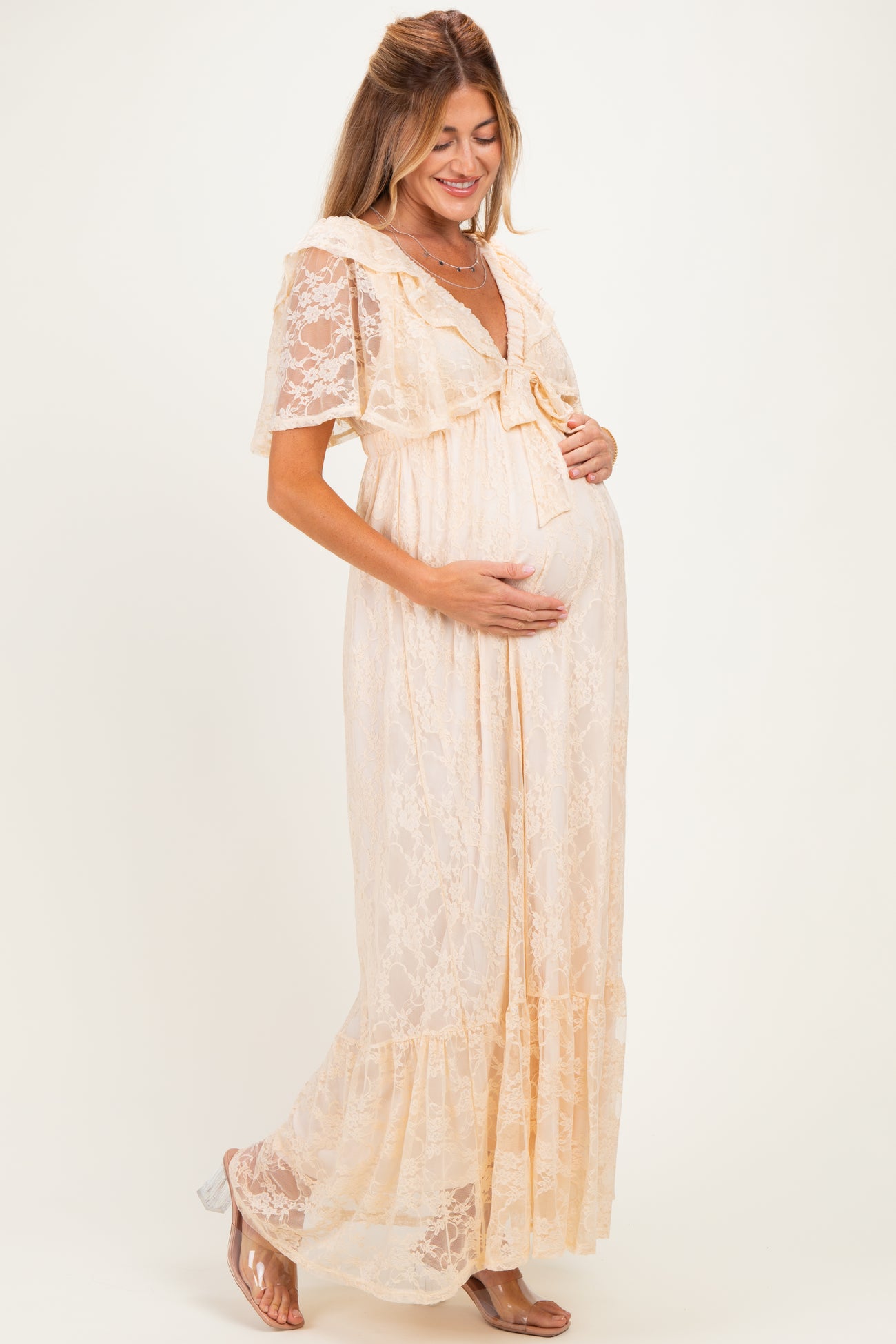 woman wearing a light peach lace maternity maxi dress with short sleeves and a bow detail, gently cradling her baby bump
