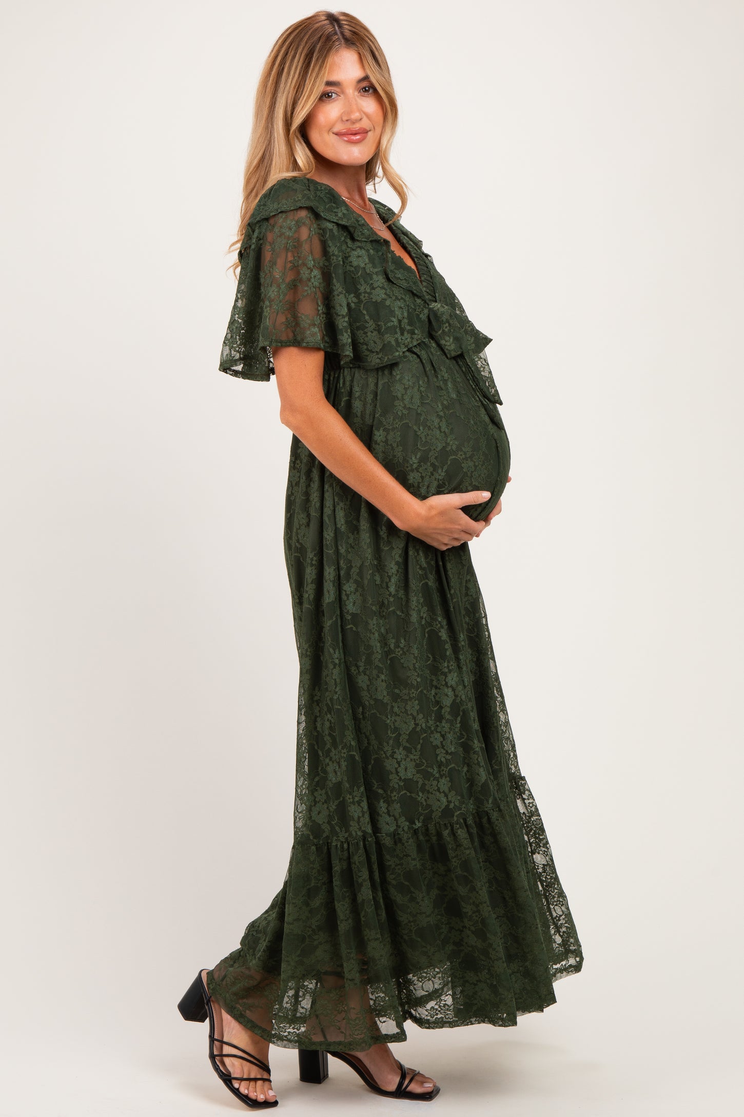 woman in a dark green lace maternity dress with flounced sleeves, holding her baby bump while posing sideways