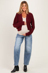 woman wearing a maroon cropped cardigan and light blue relaxed jeans, holding her baby bump in a beige top