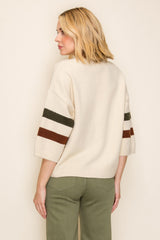 woman wearing a cream oversized sweater with brown and green stripes on the sleeves, styled with olive green pants