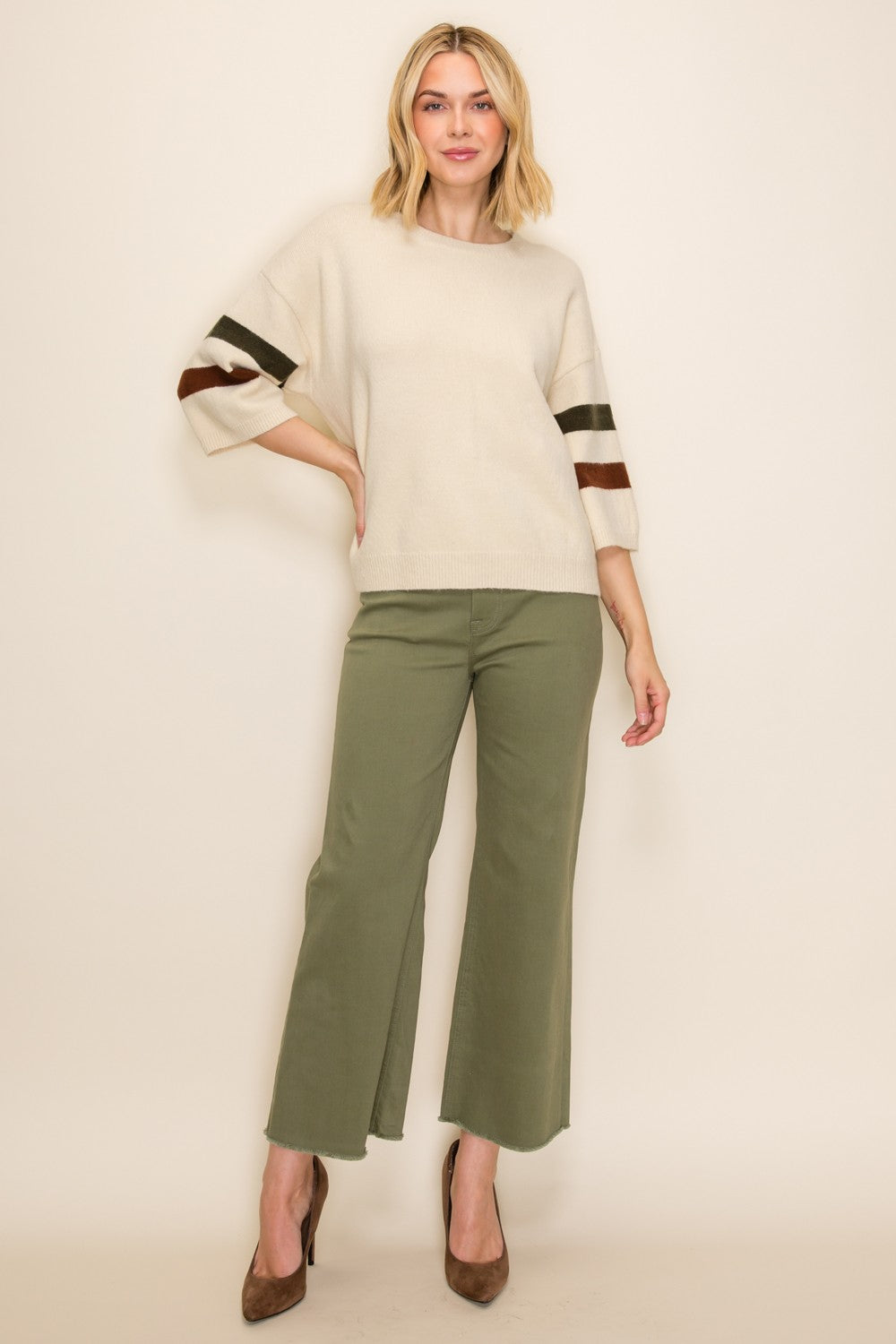 woman wearing a cream oversized sweater with striped sleeves and olive green wide-leg pants, standing in high heels