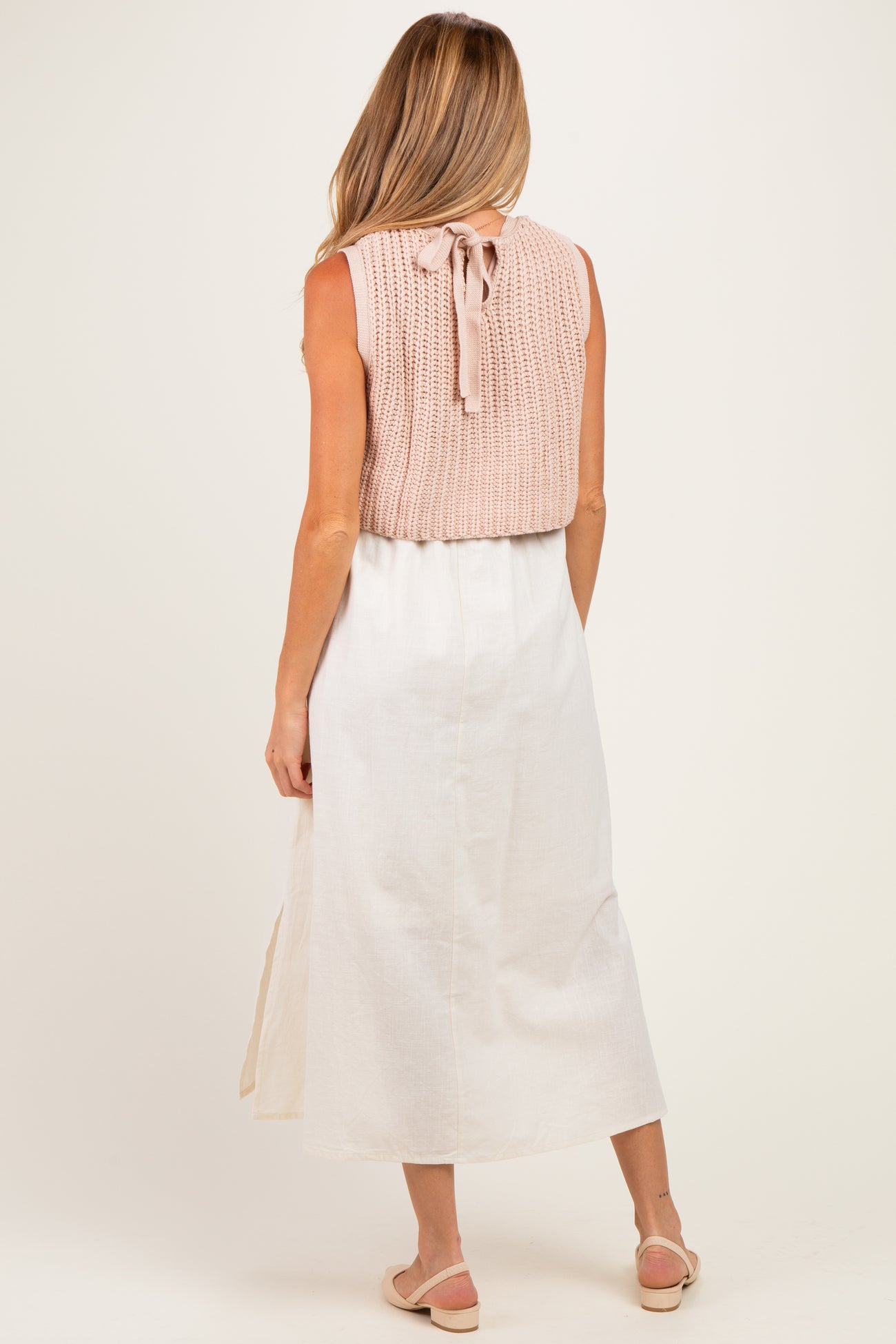 woman wearing a light pink knitted maternity top with a tie at the back and a white maxi skirt, standing with her back to the camera