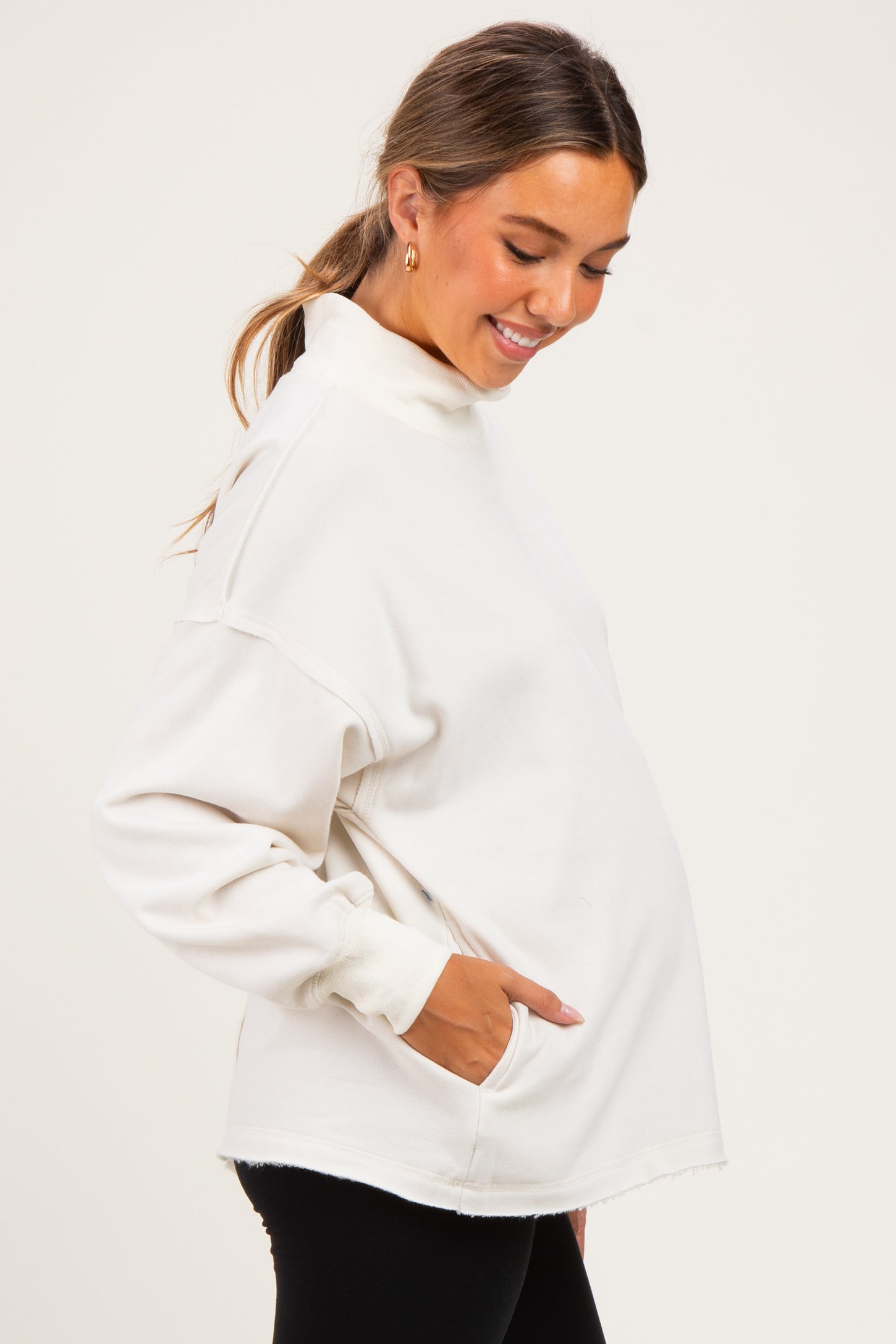 woman wearing a cream maternity pullover sweatshirt with pockets, smiling and showing her baby bump