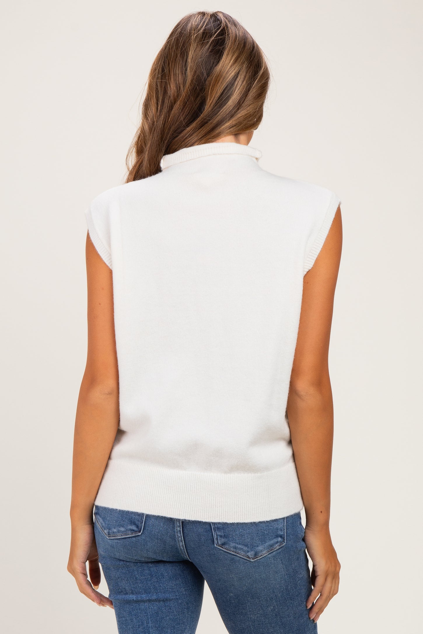 woman wearing a white sleeveless maternity sweater with a high neckline, styled with blue jeans