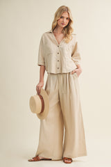 woman wearing a beige button-up short sleeve top and wide-leg pants, holding a straw hat