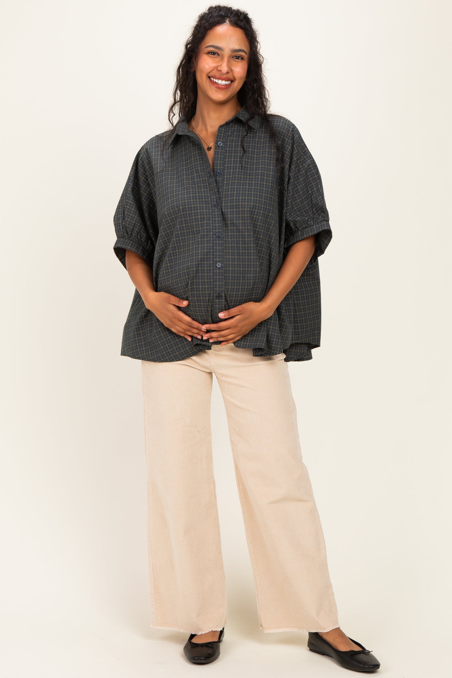 woman wearing a dark green plaid maternity shirt and beige wide-leg pants, cradling her baby bump