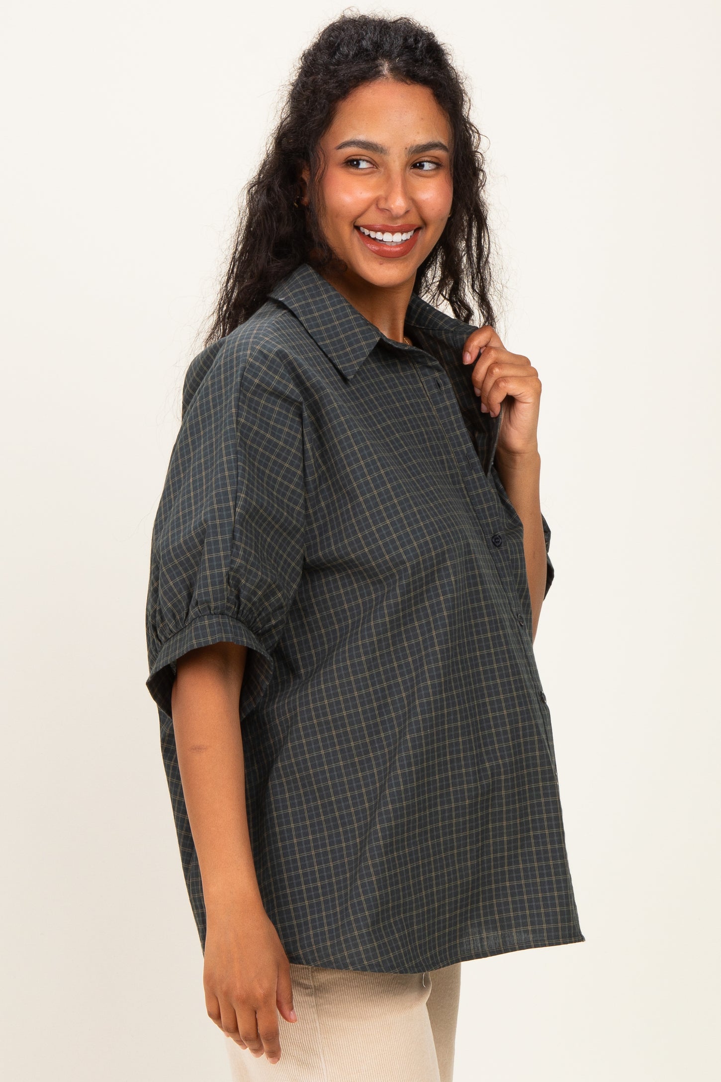 woman wearing a dark green checked maternity shirt with a relaxed fit and short sleeves, smiling while holding the collar