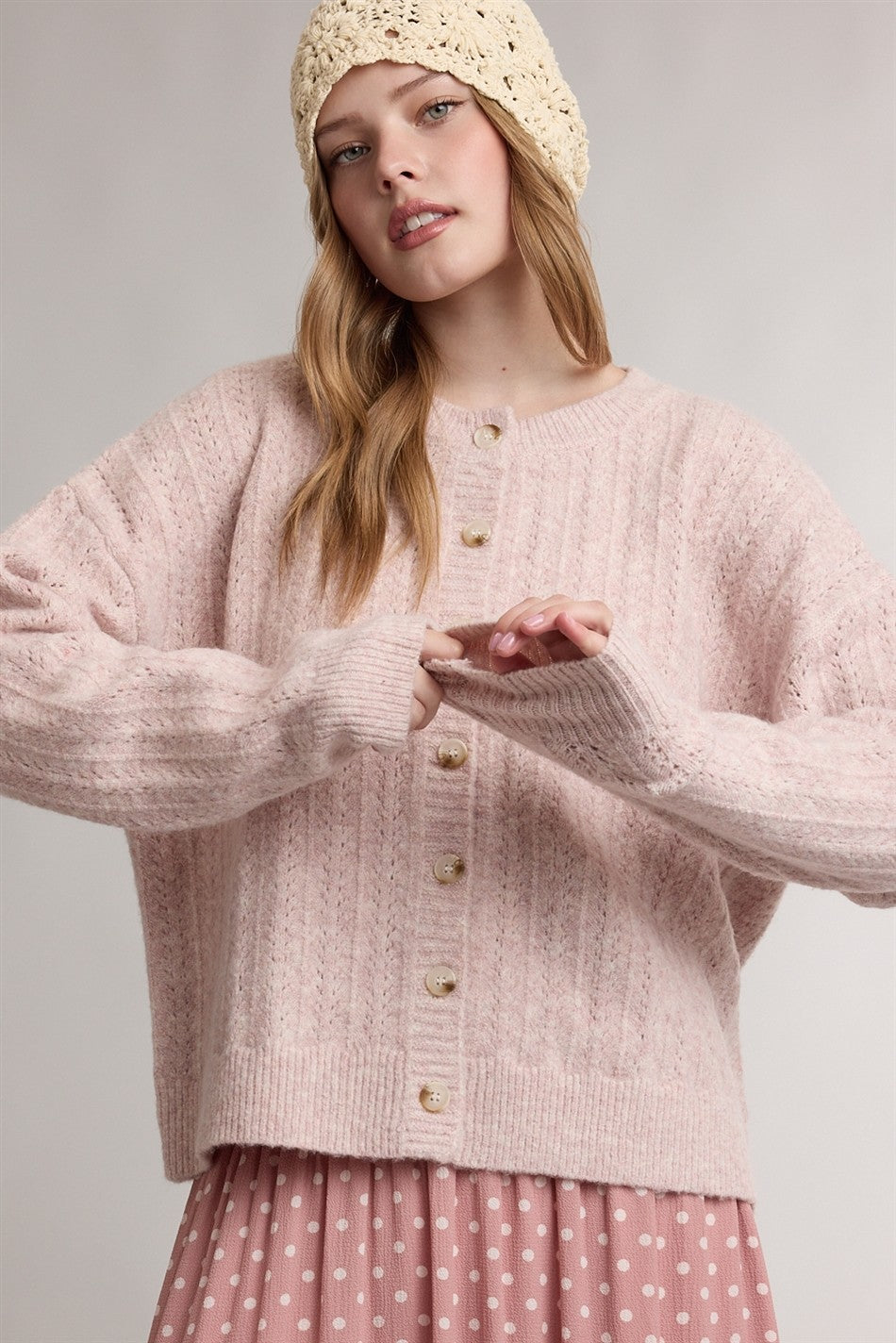 woman wearing a light pink oversized cardigan with buttons and a ribbed texture, styled with a pink polka dot skirt and a beige knit beanie