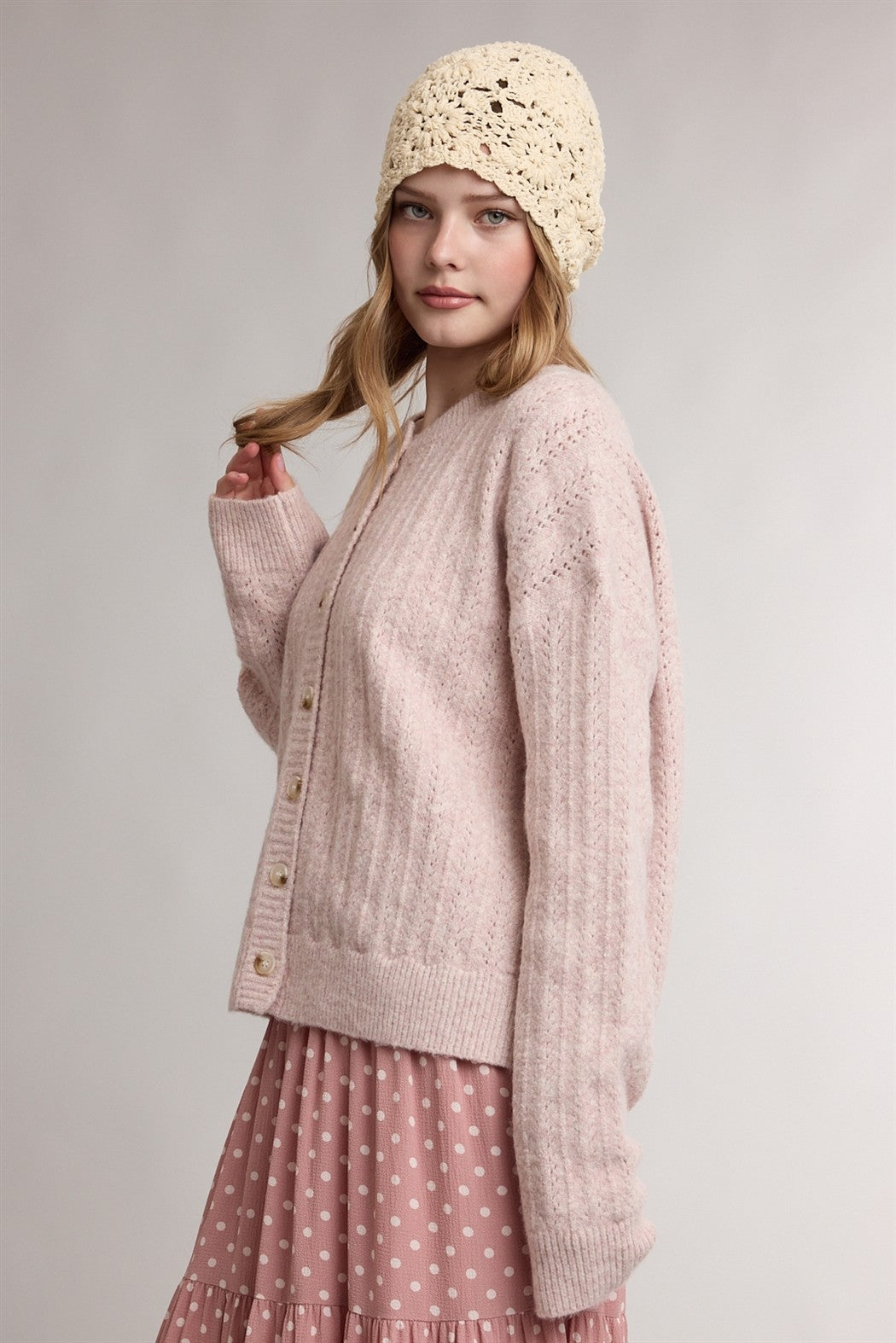 woman wearing a light pink knitted cardigan with buttons and a cream crochet beanie, styled with a polka dot pink skirt