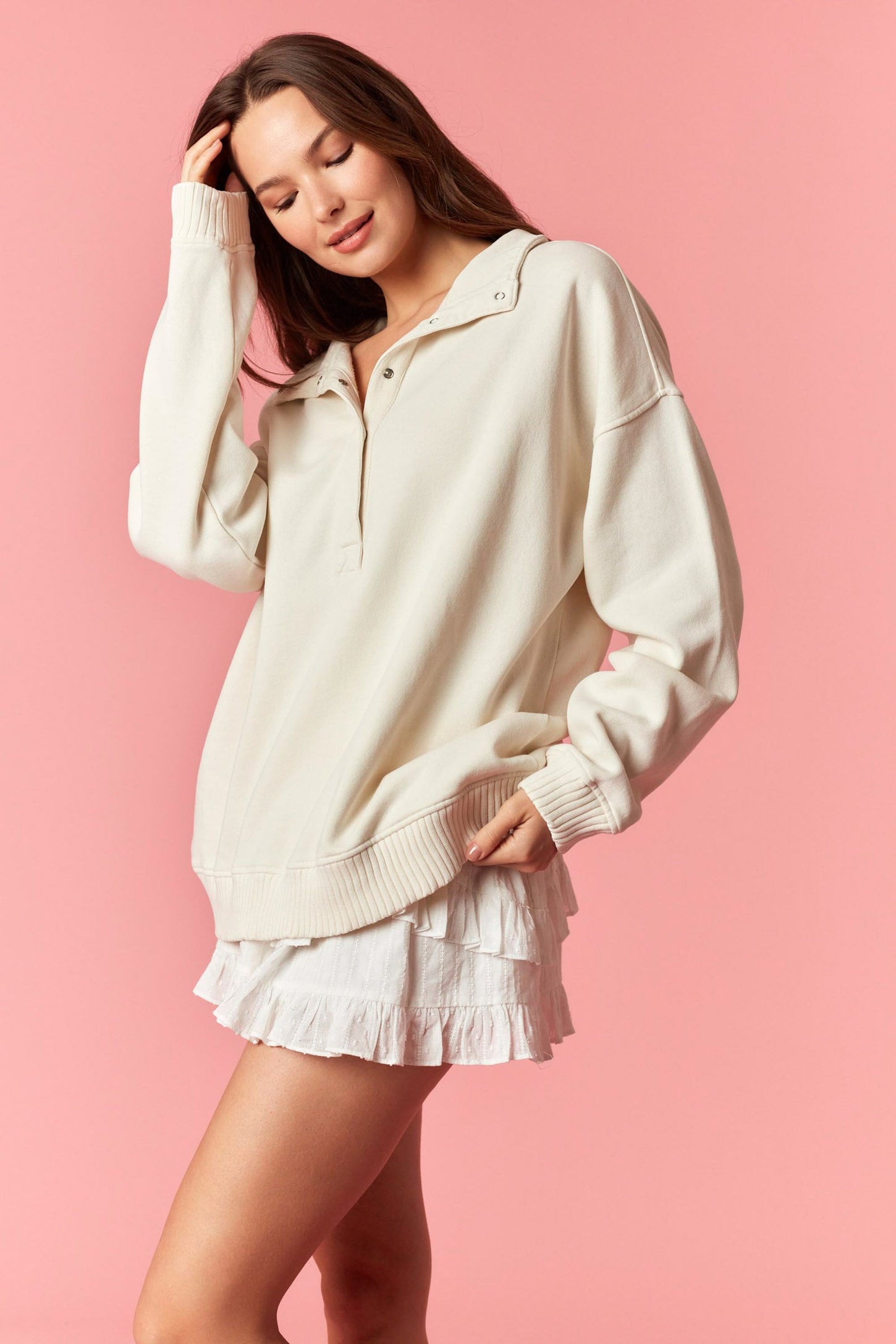 woman wearing a cream oversized quarter-zip sweatshirt with a layered ruffle skirt against a pink background