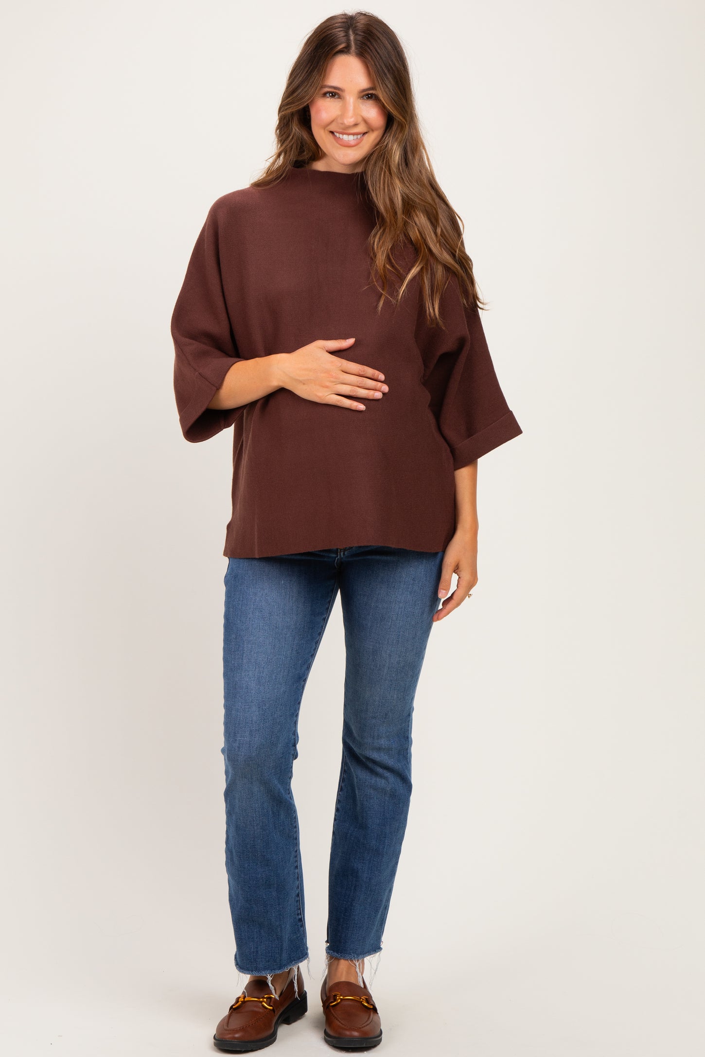 woman wearing a brown maternity top with three-quarter sleeves and holding her baby bump, paired with blue skinny jeans
