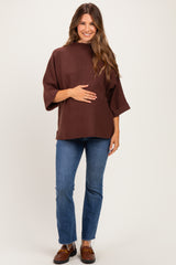 woman wearing a brown maternity top with three-quarter sleeves and holding her baby bump, paired with blue skinny jeans