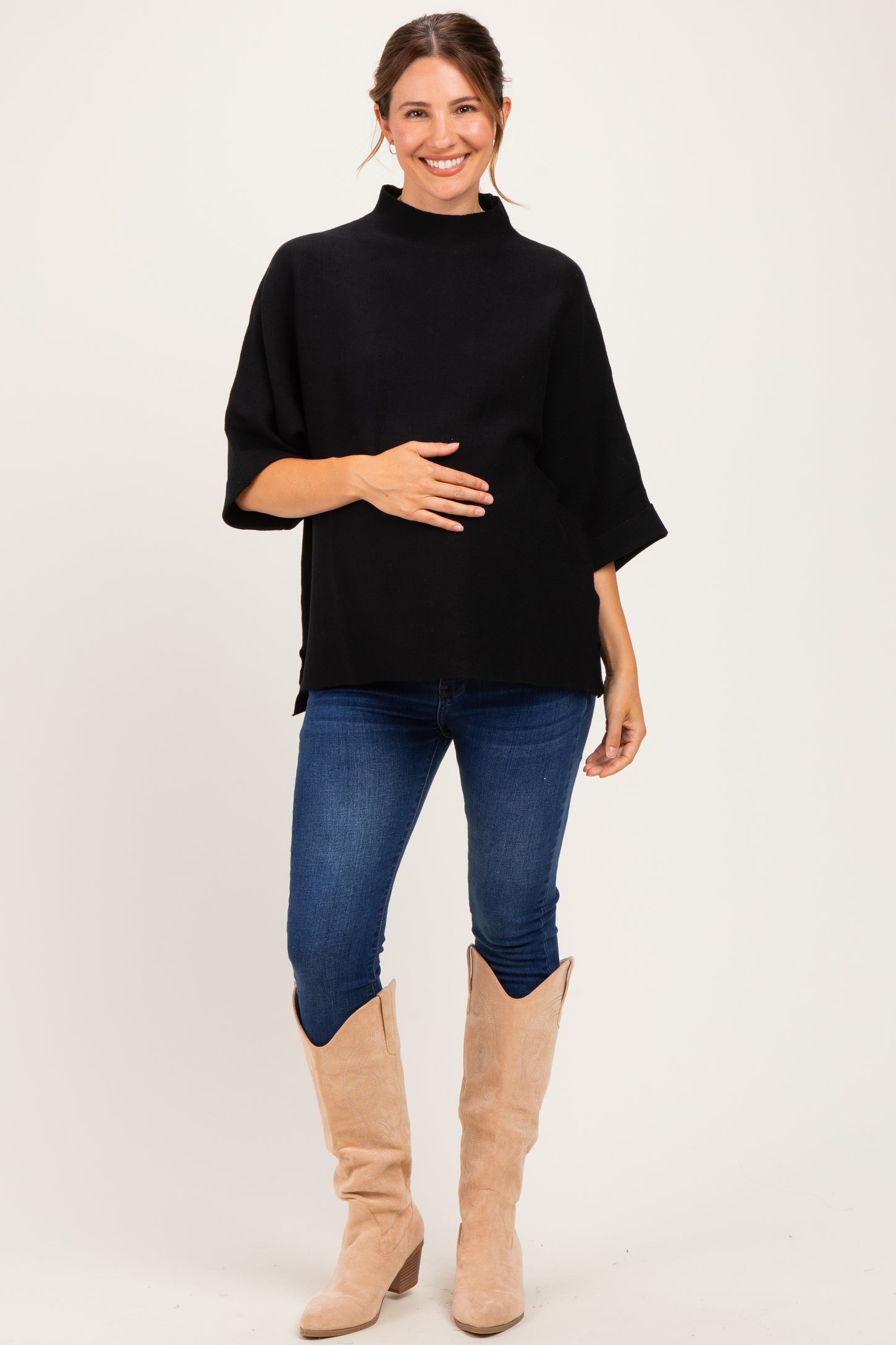 woman wearing a black short sleeve maternity top and holding her baby bump with blue jeans and beige boots