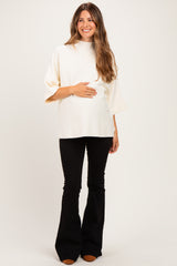 woman wearing a cream relaxed fit maternity top and black flared pants, gently holding her baby bump