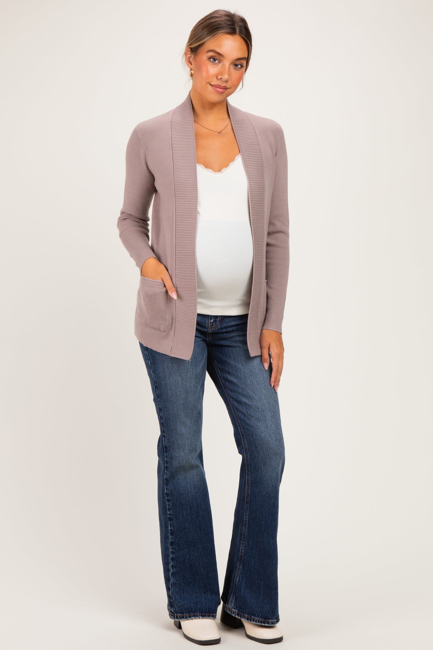 woman wearing a light mauve maternity cardigan over a white top and blue flared jeans, showing her baby bump
