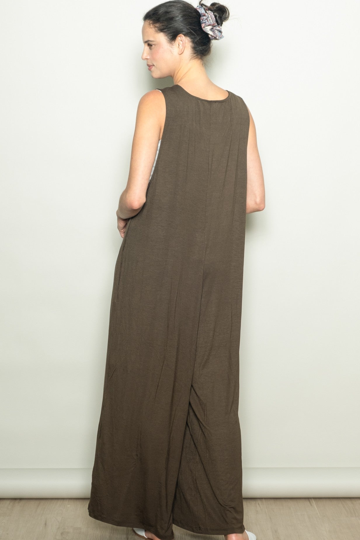 woman wearing a long brown sleeveless maxi dress, standing with her back to the camera and hands in pockets