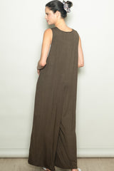 woman wearing a long brown sleeveless maxi dress, standing with her back to the camera and hands in pockets