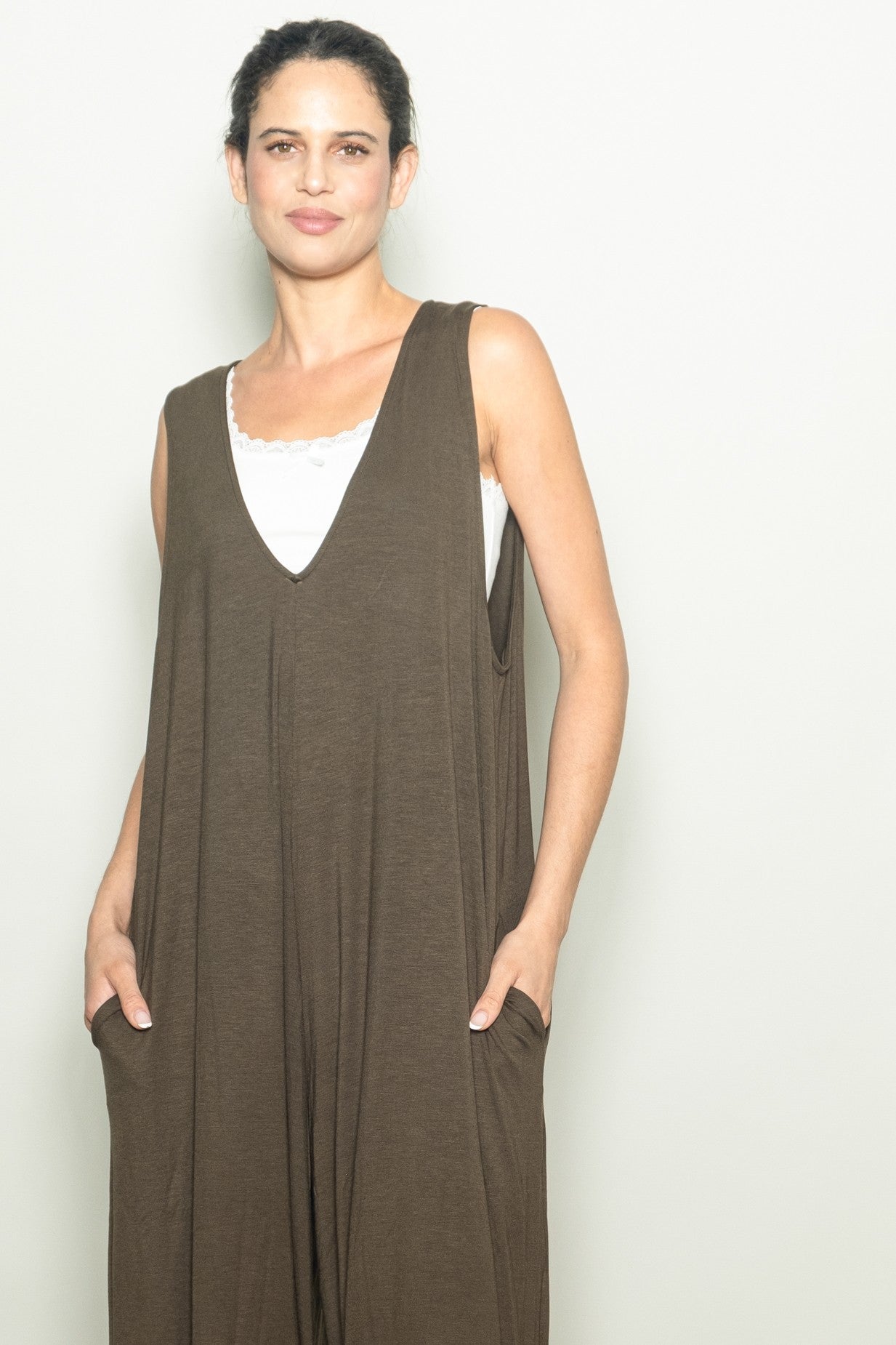 woman wearing a brown sleeveless jumpsuit with a v-neck and side pockets, styled over a white tank top