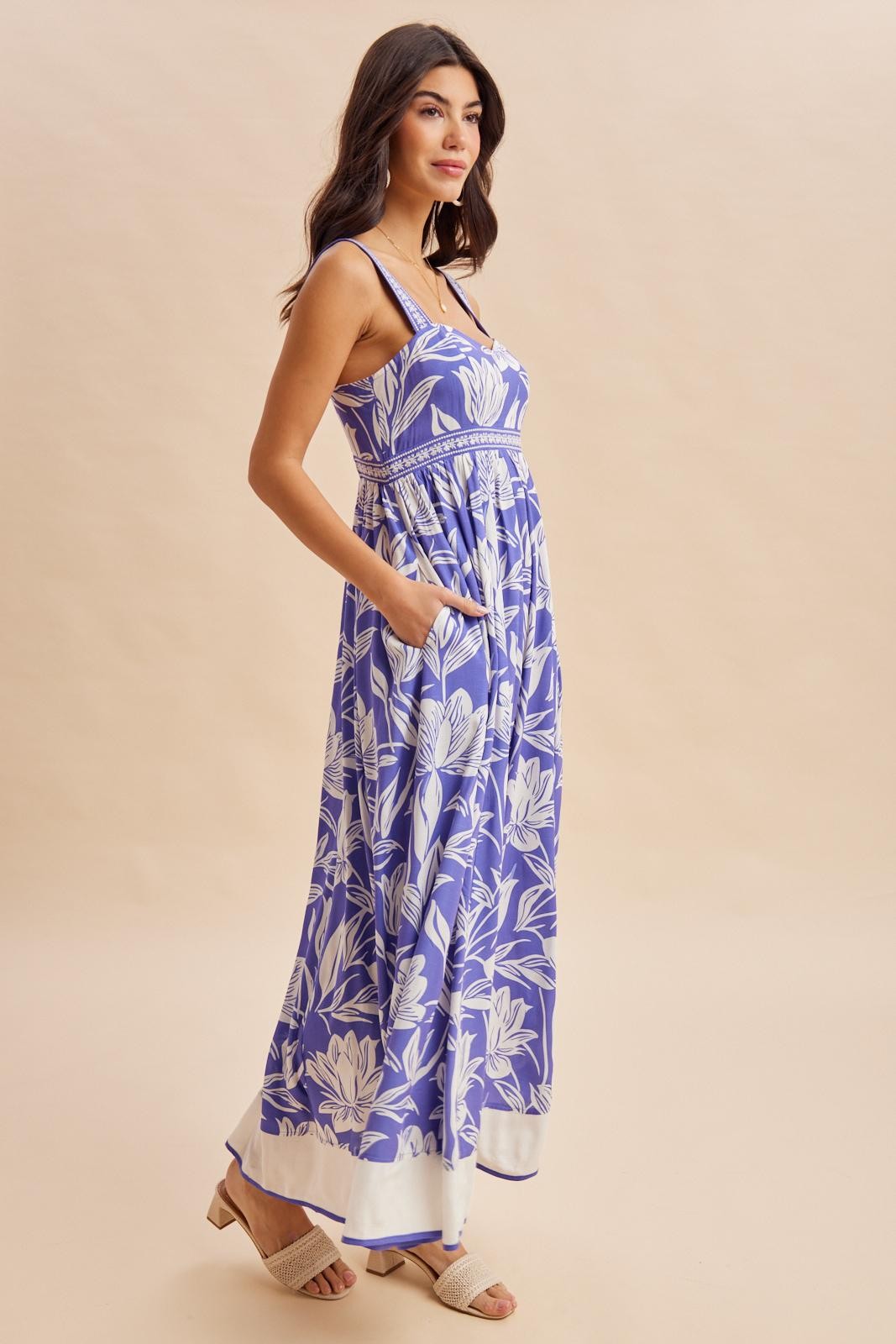 woman wearing a purple floral maxi dress with pockets and thin straps, standing against a neutral background