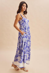 woman wearing a purple floral maxi dress with pockets and thin straps, standing against a neutral background