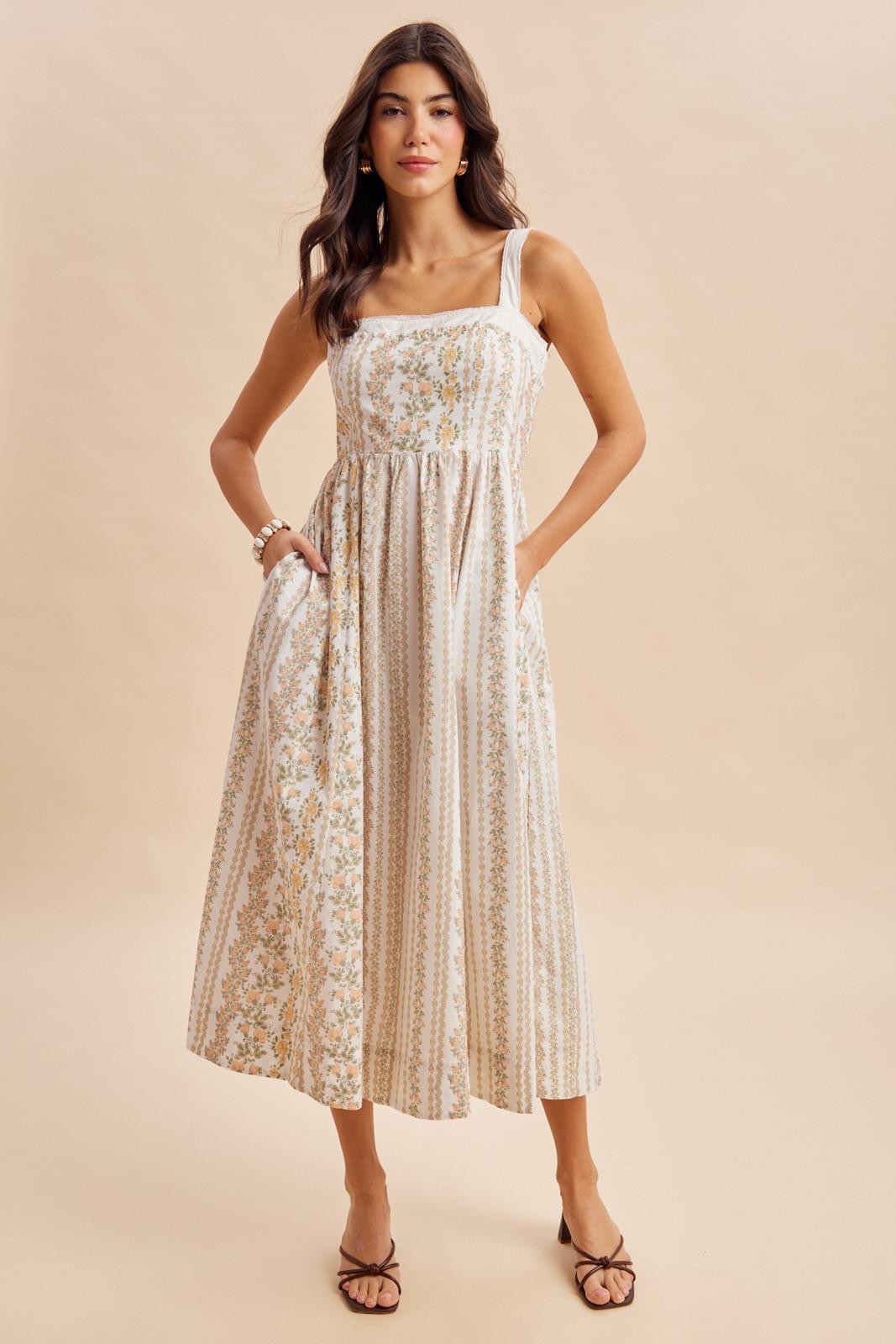 woman wearing a cream maxi dress with floral patterns and square neckline, standing with hands on hips