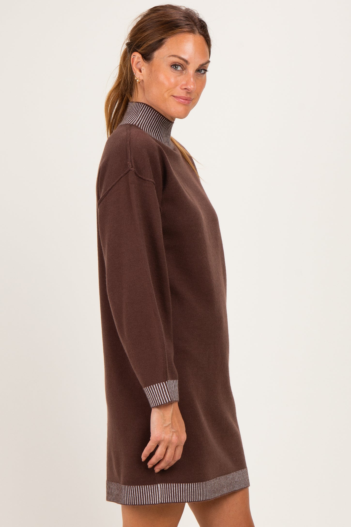 woman wearing a brown knit dress with a high neck and striped cuffs, standing sideways against a plain background