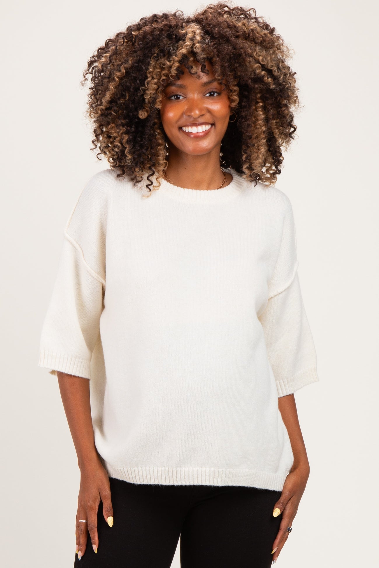woman wearing a cream oversized maternity sweater with short sleeves and a relaxed fit, smiling and showing her baby bump