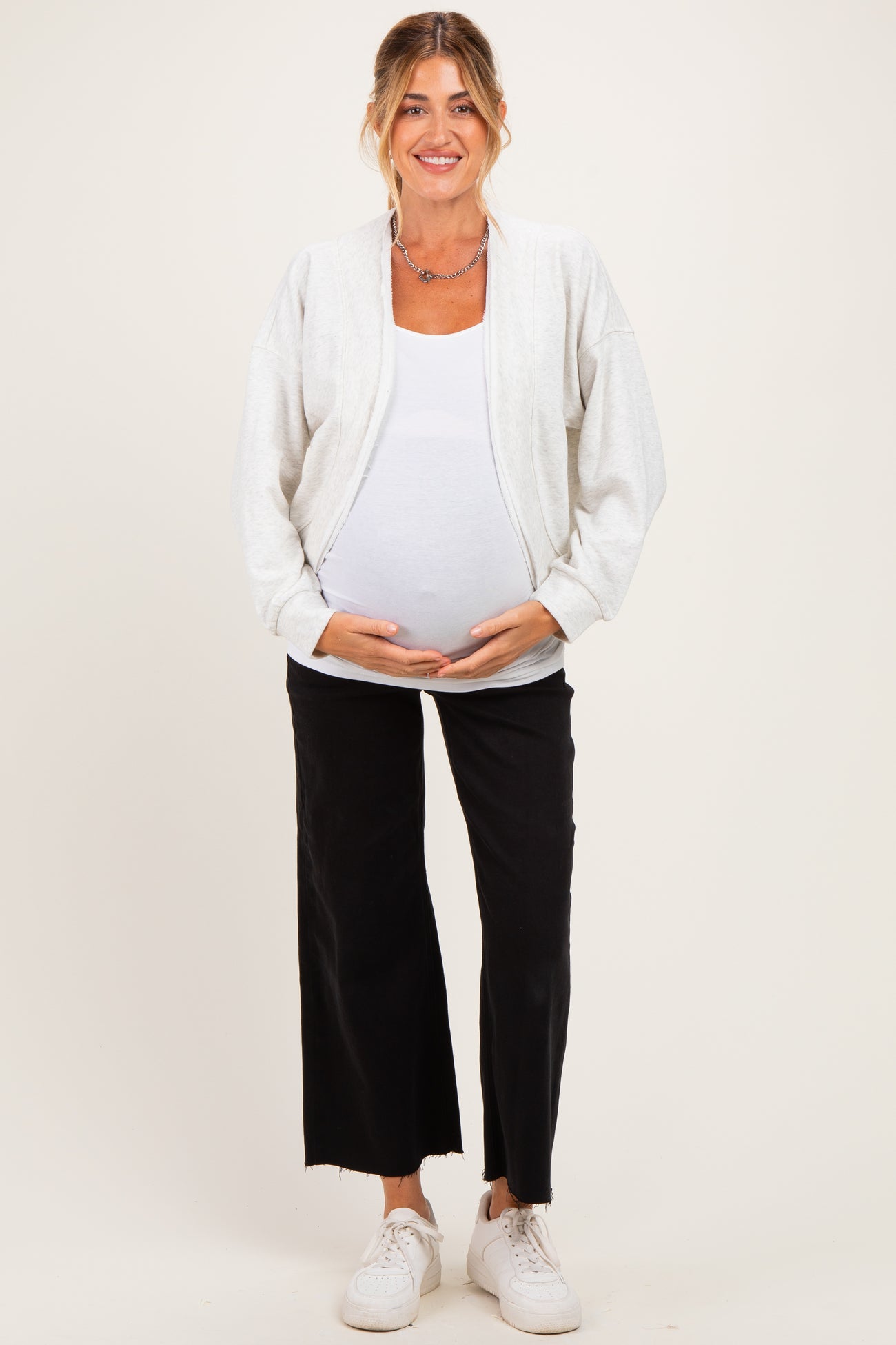 woman wearing a light gray maternity zip-up jacket over a white tank top and black wide-leg pants, showing her baby bump