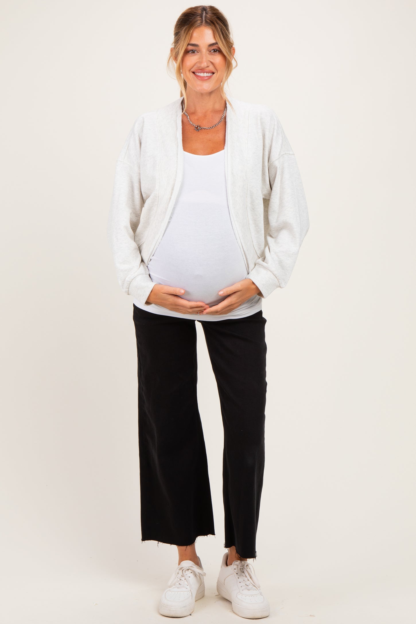 woman wearing a light gray maternity zip-up jacket over a white tank top and black wide-leg pants, showing her baby bump