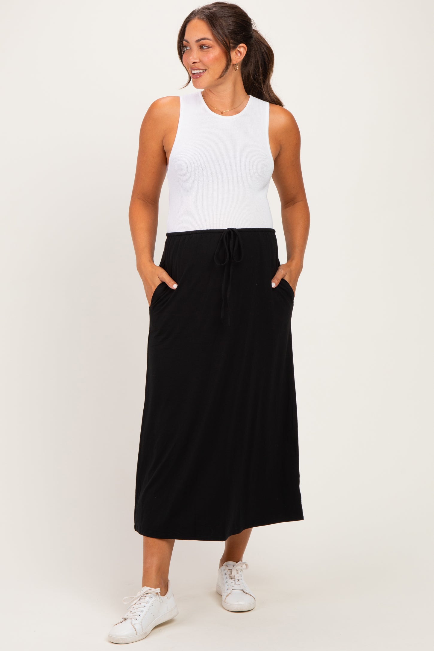 woman wearing a white ribbed tank top and a black maternity maxi dress with pockets and a drawstring waist