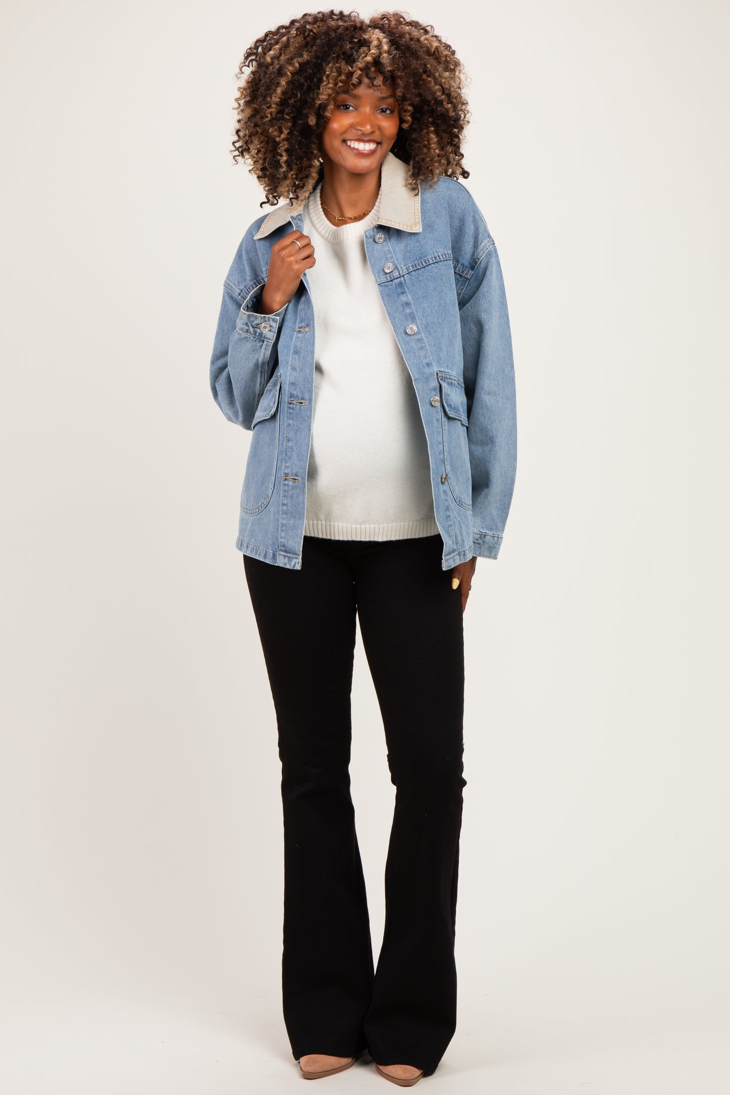 woman wearing a light blue denim jacket over a white sweater, showing her baby bump in black flared maternity pants