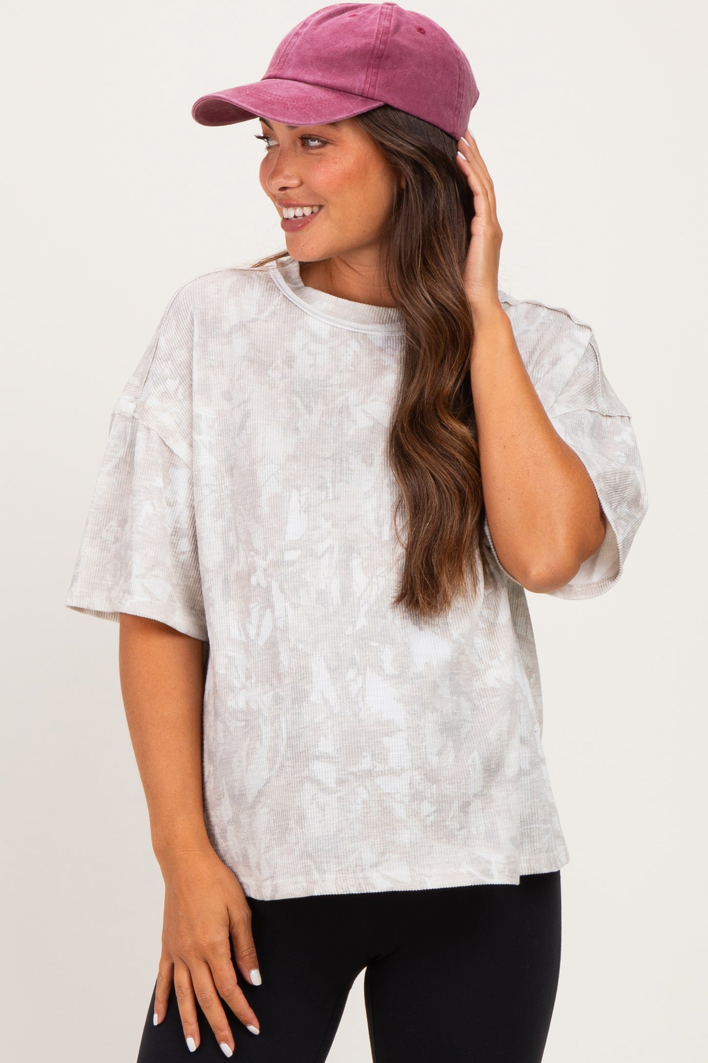 woman wearing a light gray oversized maternity top with a subtle pattern and a pink baseball cap, smiling and touching her hair