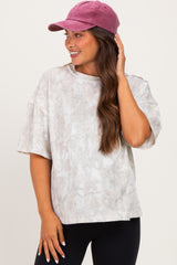 woman wearing a light gray oversized maternity top with a subtle pattern and a pink baseball cap, smiling and touching her hair