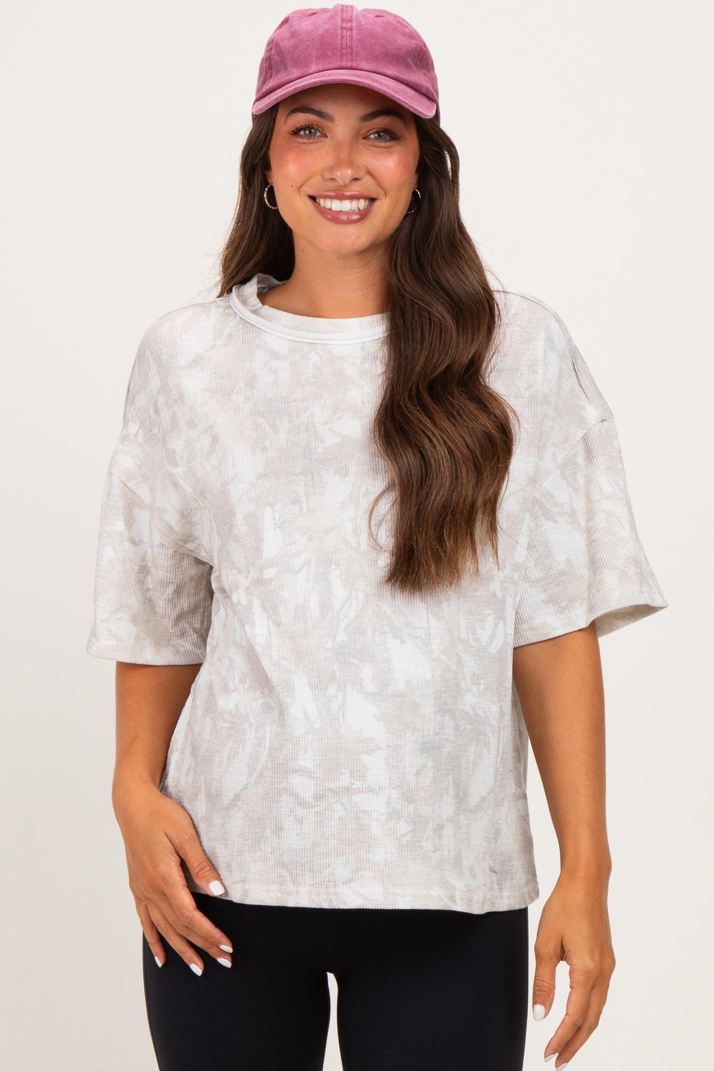 woman wearing a light gray oversized maternity top with a subtle pattern and a pink baseball cap, smiling at the camera